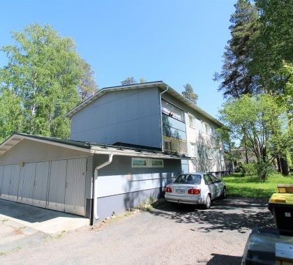 Aurinkoinen näkymä vaaleansiniseen kaksikerroksiseen asuinkerrostaloon ja sen autotalliin vehreän pihan ympäröimänä. Kuva esittää huolellisesti hoidettua asuinkiinteistöä ja tyypillistä isännöintikohdetta.