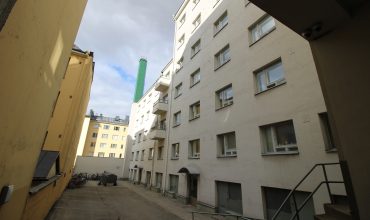 Korkea, vaaleasävyinen asuinkerrostalo ja sen asfaltoitu sisäpiha kaupunkiympäristössä. Kuvassa näkyy taloyhtiön rakennuksen huollettu julkisivu.