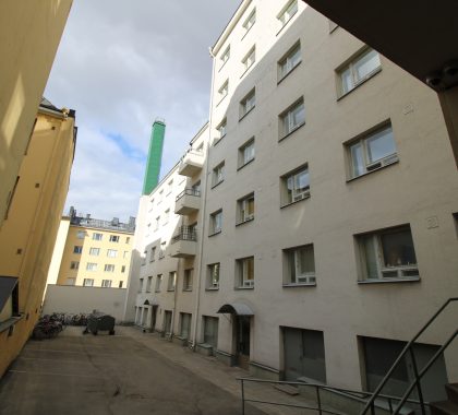 Korkea, vaaleasävyinen asuinkerrostalo ja sen asfaltoitu sisäpiha kaupunkiympäristössä. Kuvassa näkyy taloyhtiön rakennuksen huollettu julkisivu.